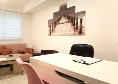 Escritorio blanco de consulta de psicología con un portapapeles y, al fondo, un sofá rosa y un cuadro decorativo de un puente de madera en la pared.